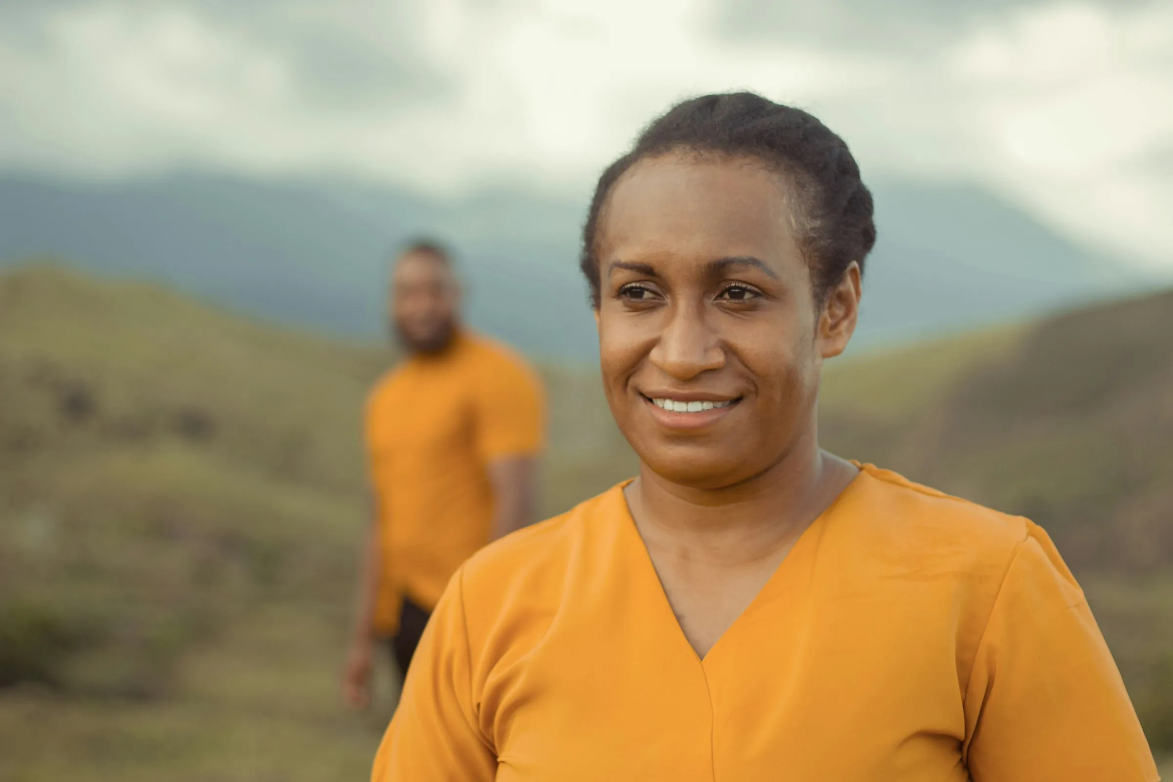 A smiling woman in an orange top outdoors, with green hills behind her